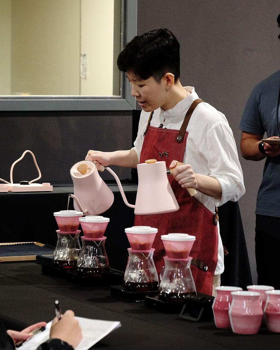 Peace brewing at the USBrC. She holds two pink gooseneck kettles in either hand, brewing in front of four pink coffee droppers. In the foreground, four pink 2-Way Cups sit on the table.